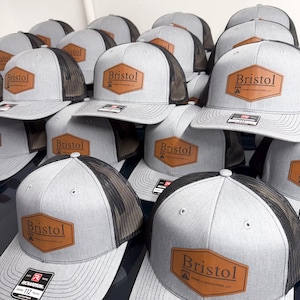 May include: A collection of gray trucker hats with black mesh backs and flat brims. Each hat features a brown hexagonal leather patch on the front with the text Bristol Prime Contractors LLC printed in black. A black sticker is on the brim.