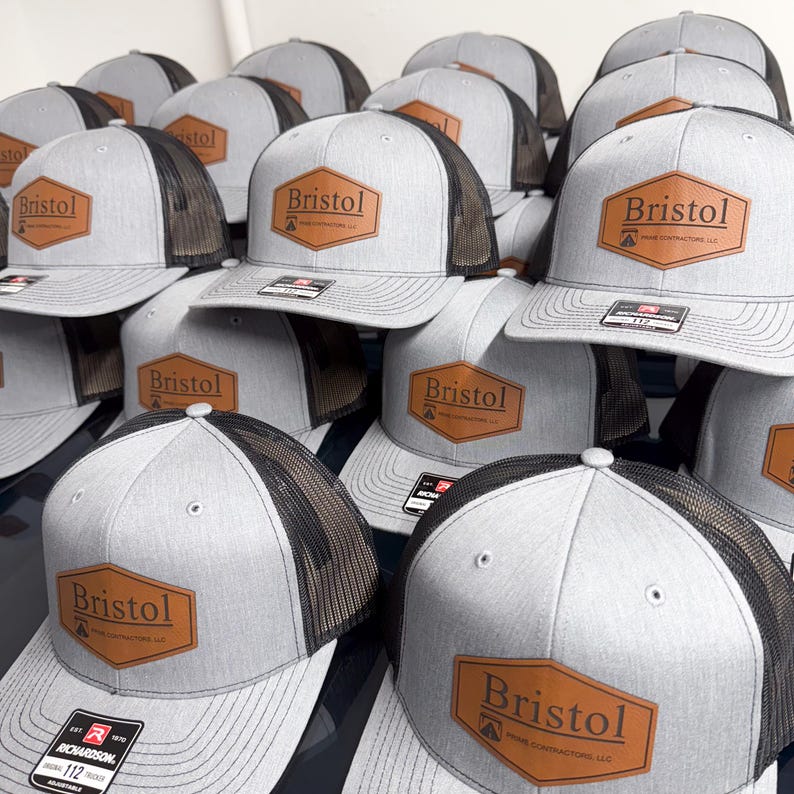 May include: A collection of gray trucker hats with black mesh backs and flat brims. Each hat features a brown hexagonal leather patch on the front with the text Bristol Prime Contractors LLC printed in black. A black sticker is on the brim.