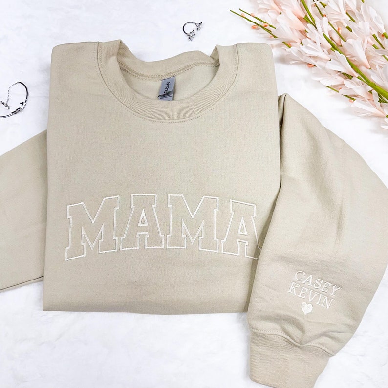 May include: A beige sweatshirt with the word "MAMA" embroidered in white on the front. The left sleeve has the names "CASEY" and "KEVIN" embroidered in white with a heart symbol.
