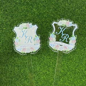Op de afbeelding: Twee heldere acryl bruiloft decoratiestukken met een schildvorm, met bloemstukken in roze, blauw en wit. Het ontwerp bevat de initialen "K R" en de datum "15 januari 2025". Elk stuk heeft een heldere steel.