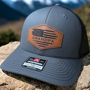 May include: A gray and black trucker hat with a brown leather patch that says "Grandpa" and features a distressed American flag design. The hat is adjustable and has a Richardson 112 logo.