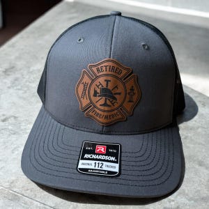 May include: A charcoal gray trucker hat with a black mesh back and a brown leather patch on the front. The patch features a fire department emblem with the text RETIRED FIRE/MEDIC, a helmet, ladder, and hydrant. A black sticker on the brim reads RICHARDSON 112.