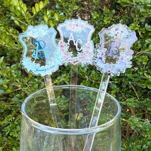 Op de afbeelding: Drie heldere acryl roerstaafjes met bloemen- en initiaalontwerpen. Elk roerstaafje heeft een andere initiaal, omringd door bloemen en data. De roerstaafjes zitten in een glas, met een groene, bladerrijke achtergrond.