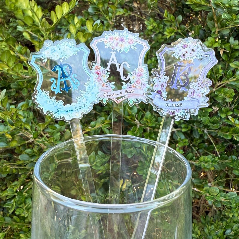 Op de afbeelding: Drie heldere acryl roerstaafjes met bloemen- en initiaalontwerpen. Elk roerstaafje heeft een andere initiaal, omringd door bloemen en data. De roerstaafjes zitten in een glas, met een groene, bladerrijke achtergrond.