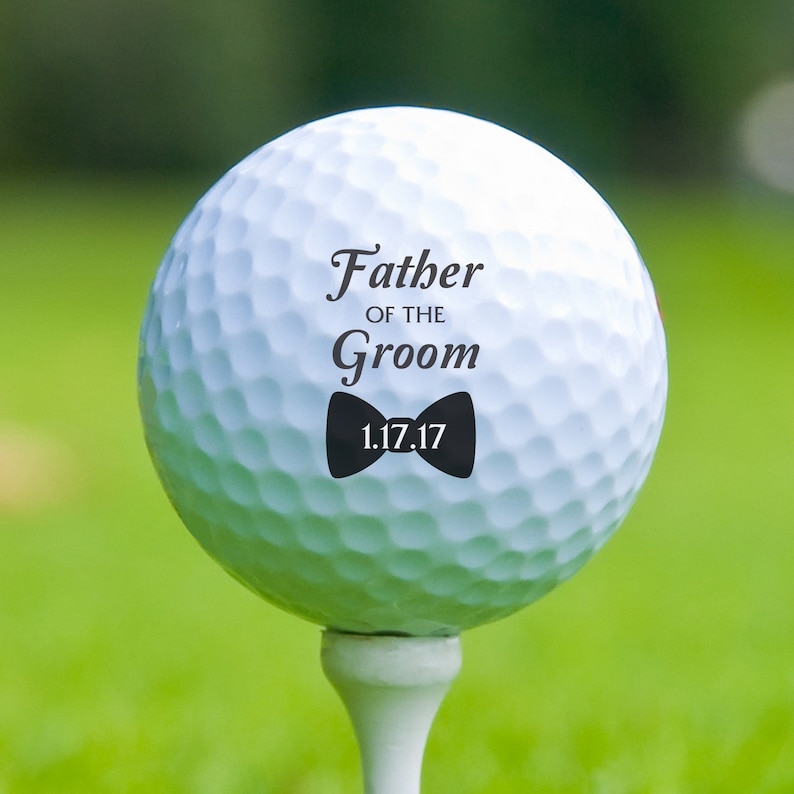 K&ouml;nnte beinhalten: Ein wei&szlig;er Golfball auf einem Tee mit dem Text "Father of the Groom" und einer schwarzen Fliege sowie dem Datum "1.17.17". Der Ball befindet sich auf einem gr&uuml;nen Golfplatz.