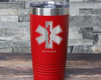 May include: A red insulated tumbler with a silver lid. The tumbler features a silver Star of Life emblem with a red staff and snake, and the name "Bob Sanders" below the emblem. The tumbler has a ridged design near the base.