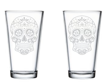 May include: Two clear glass pint glasses featuring a white sugar skull design. The skull is decorated with floral patterns.