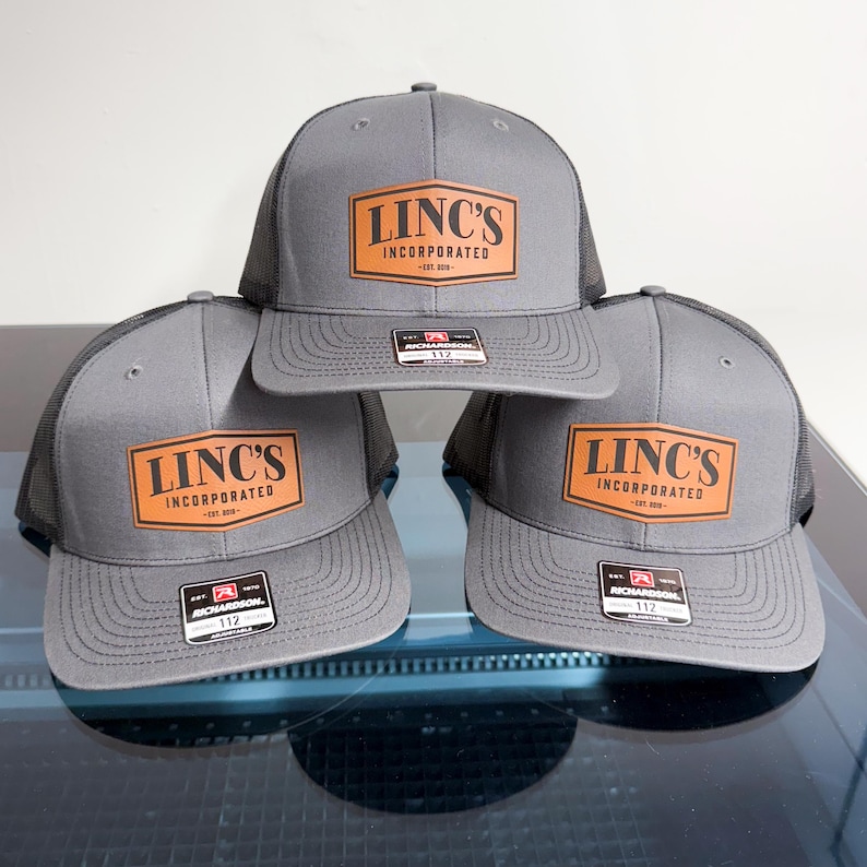 May include: Three gray trucker hats with black mesh backs and flat brims. Each hat features a tan leather-like patch on the front with the black text LINC'S INCORPORATED EST. 2019. A small black and red Richardson 112 adjustable sticker is on each brim.