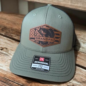 May include: Olive green trucker hat with a black mesh back. The front features a brown leather patch with a fish graphic and the word "GRANDPA". The hat includes a Richardson 112 Trucker tag.