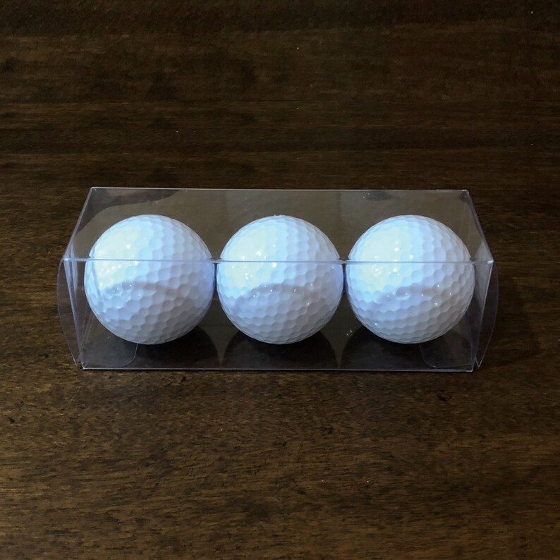 K&ouml;nnte beinhalten: Drei wei&szlig;e Golfb&auml;lle mit geriffelten Oberfl&auml;chen werden in einem transparenten Kunststoffbeh&auml;lter pr&auml;sentiert. Die Golfb&auml;lle sind nebeneinander in dem rechteckigen Beh&auml;lter angeordnet. Der Hintergrund ist eine dunkelbraune Holzoberfl&auml;che.