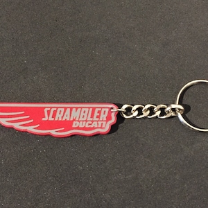 Op de afbeelding: Rode en zilveren sleutelhanger met een vleugelontwerp en de tekst "Scrambler Ducati".