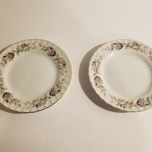 May include: Two white ceramic salad plates with a floral design featuring pink roses and green leaves. The plates have a gold trim around the edge.