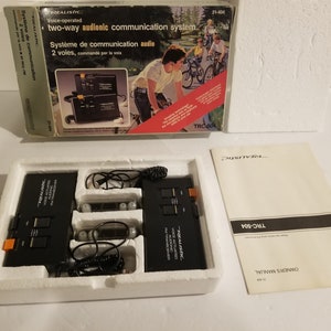 May include: A Realistic TRC-504 voice-operated two-way audionic communication system in its original packaging. The box features a red and white design with an illustration of people riding bicycles. The system includes two black units with earpieces and a cord.