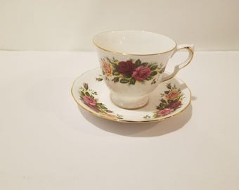 Pode incluir: Uma chávena de chá e um pires brancos com um padrão floral de rosas cor-de-rosa, amarelas e verdes. A chávena e o pires têm uma guarnição dourada.
