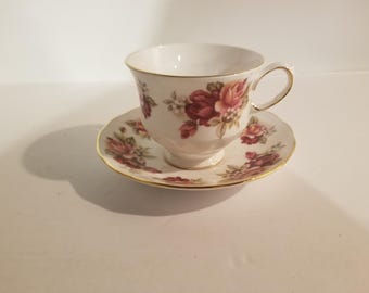 Puede incluir: Una taza de té y un platillo blancos con un patrón floral con rosas rosas y rojas. La taza y el platillo tienen un borde dorado.