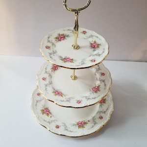Op de afbeelding: Een drielaags witte porseleinen taartstandaard met een gouden rand en een bloemenmotief. De standaard heeft een gouden handvat en is perfect om desserts of snacks te serveren.