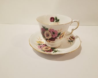 May include: A white teacup and saucer set with a floral design. The teacup and saucer have a gold trim. The floral design features pink, purple, and yellow flowers with green leaves. The teacup has a curved handle.