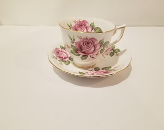 May include: A white teacup and saucer with a pink rose floral pattern. The cup has a gold rim and the saucer has a gold trim.