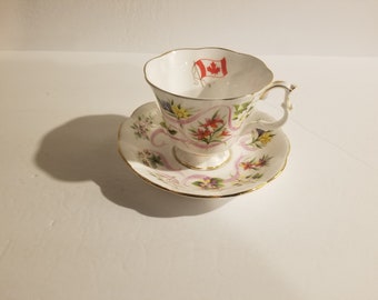 May include: A white teacup and saucer with a floral design and a Canadian flag. The teacup has a gold rim and the saucer has a gold rim and a gold band around the edge.