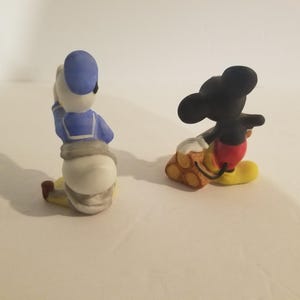 Puede incluir: Dos figuritas de cer&aacute;mica de Mickey Mouse y Donald Duck. Mickey Mouse lleva pantalones cortos rojos y zapatos amarillos y lleva una pila de troncos. Donald Duck lleva una camisa de marinero azul y pantalones blancos.