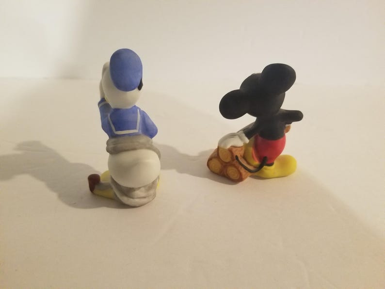 Puede incluir: Dos figuritas de cer&aacute;mica de Mickey Mouse y Donald Duck. Mickey Mouse lleva pantalones cortos rojos y zapatos amarillos y lleva una pila de troncos. Donald Duck lleva una camisa de marinero azul y pantalones blancos.