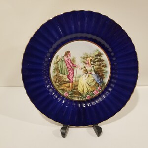 May include: A blue and white porcelain plate with a gold rim. The plate features a romantic scene of a couple in a garden setting.