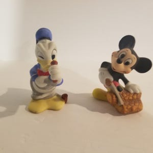 Puede incluir: Dos figuritas de cer&aacute;mica vintage de Mickey Mouse y Donald Duck. Mickey Mouse lleva pantalones cortos rojos y sostiene un objeto marr&oacute;n. Donald Duck lleva un gorro de marinero azul y una pajarita roja.