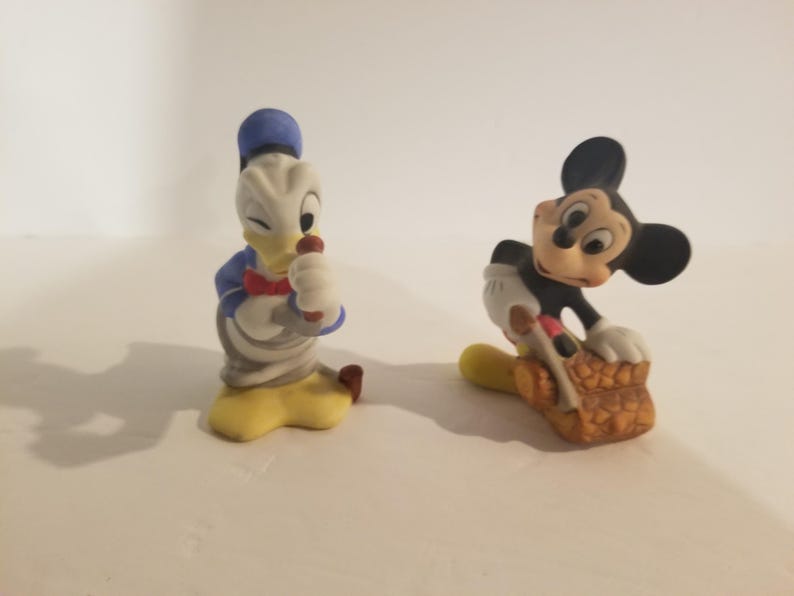 Puede incluir: Dos figuritas de cer&aacute;mica vintage de Mickey Mouse y Donald Duck. Mickey Mouse lleva pantalones cortos rojos y sostiene un objeto marr&oacute;n. Donald Duck lleva un gorro de marinero azul y una pajarita roja.
