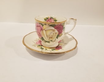 May include: A white teacup and saucer set with a floral design featuring pink and white roses. The cup and saucer have a gold trim.