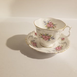 Puede incluir: Un juego de taza y platillo de té blanco con un patrón floral de rosas rosas y nomeolvides azules. El juego tiene un borde dorado.