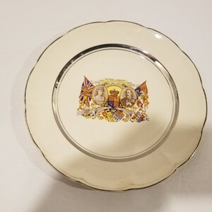 Puede incluir: Un plato de cerámica blanco con un borde plateado y un diseño conmemorativo para el Jubileo de Plata del rey Jorge V y la reina María en 1935. El diseño presenta retratos del rey y la reina, el escudo de armas real y dos banderas.