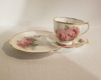 May include: A white teacup and saucer set with a pink rose floral pattern. The saucer has a scalloped edge and a gold trim. The teacup has a handle and a gold trim around the rim.