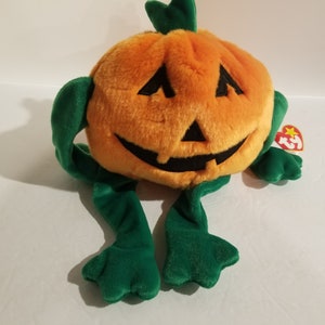 May include: Orange and green plush pumpkin with a smiling jack-o'-lantern face. The pumpkin has long green arms and legs. The plush toy has a Ty Beanie Baby tag attached.