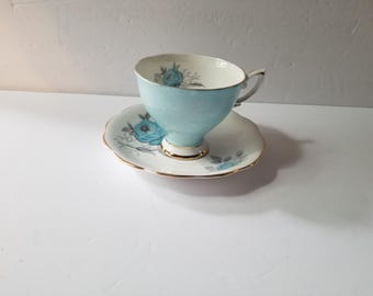May include: A vintage teacup and saucer set in light blue and white. The teacup has a curved shape and a handle, with a gold trim. The saucer has a scalloped edge and gold trim. Both pieces are decorated with blue floral patterns.