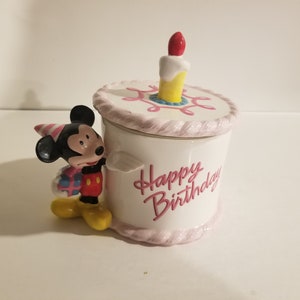 May include: White ceramic cookie jar shaped like a birthday cake with a pink frosting design. The lid is a separate piece and features a single lit candle. Mickey Mouse is standing next to the jar, holding a gift. The jar says "Happy Birthday" in pink lettering.