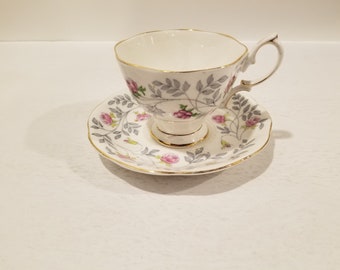 Può includere: Un set da tè bianco con un motivo floreale. La tazza e il piattino presentano un delicato disegno di rose rosa e foglie grigie su uno sfondo bianco con un bordo dorato.