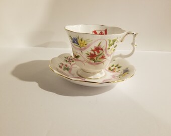 May include: A white teacup and saucer with a floral design featuring pink, red, yellow, and blue flowers. The cup and saucer have a gold trim and a pink ribbon with the word "Canada" written on it.