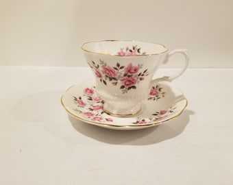 May include: A white teacup and saucer set with a pink rose floral pattern. The cup and saucer have a gold trim.
