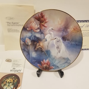 以下が含まれることがあります： 金色の縁取りのある白い磁器の皿には、ピンクと紫の水蓮のある池に立っている2羽の白いサギの絵が描かれています。皿のタイトルは「The Egrets」で、「Wings of Beauty」コレクションの一部です。皿はスタンドの上に置かれています。