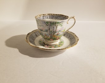 Può includere: Un set vintage di tazza e piattino da tè con un motivo floreale e una rappresentazione di betulle. La tazza e il piattino sono bianchi con un bordo dorato e un motivo grigio e bianco.