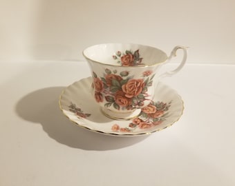 Pode incluir: Um conjunto de chávena e pires branco com um padrão floral de rosas cor-de-rosa e laranja. O conjunto tem uma borda dourada.