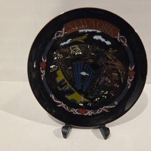 May include: Black decorative plate with a gold rim, featuring a colourful illustration of New York City and the text "NEW YORK". The plate has a decorative border with hearts and ribbons. It is displayed on a black stand.