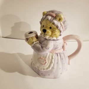 Puede incluir: Una tetera de cerámica con forma de oso que lleva un vestido lavanda y un gorro rosa. La tetera tiene un asa rosa y un pico blanco con forma de taza.