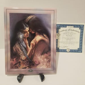 May include: A framed print depicting two women in a tender embrace, with one gently touching the other's face. The artwork features soft colors and a Native American theme. A certificate of authenticity is visible.