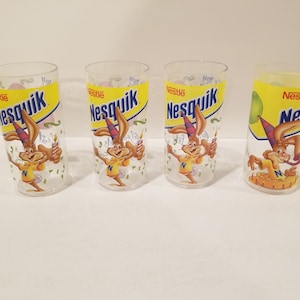 Puede incluir: Conjunto de cuatro vasos de plástico transparente Nesquik con un conejo de dibujos animados que lleva un gorro de fiesta y sostiene una vela encendida. Los vasos son amarillos con detalles azules y rojos. El texto "Nestle Nesquik" está impreso en la parte delantera de cada vaso.
