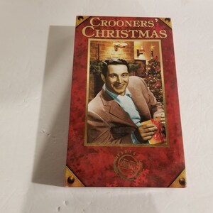 Può includere: Scatola rettangolare con il titolo "Crooners' Christmas" in lettere dorate. La copertina presenta un uomo in giacca e cravatta che tiene un regalo, con un albero di Natale e un camino sullo sfondo. La scatola ha una combinazione di colori rosso e oro.