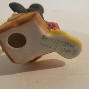 Puede incluir: Una figurita de cer&aacute;mica de Mickey Mouse sosteniendo una manzana roja. La figurita est&aacute; pintada en negro, amarillo y marr&oacute;n. La parte inferior de la figurita est&aacute; marcada con "WALT DISNEY PRODUCTIONS" en texto verde.