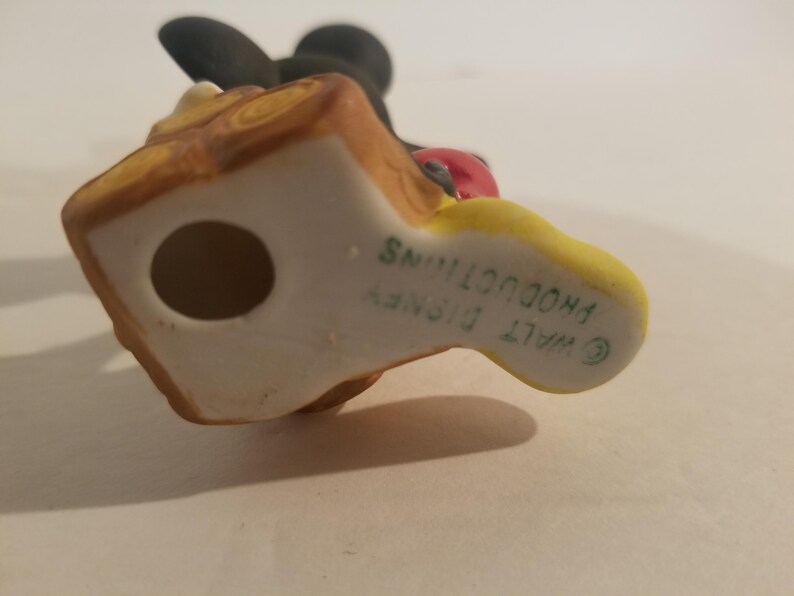 Puede incluir: Una figurita de cer&aacute;mica de Mickey Mouse sosteniendo una manzana roja. La figurita est&aacute; pintada en negro, amarillo y marr&oacute;n. La parte inferior de la figurita est&aacute; marcada con "WALT DISNEY PRODUCTIONS" en texto verde.