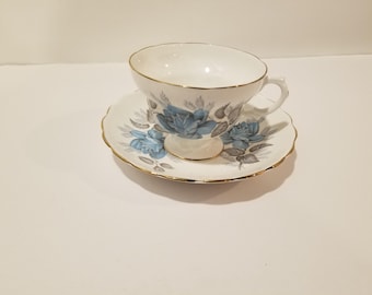 May include: A white teacup and saucer with a blue and grey floral pattern. The cup and saucer have a gold trim.