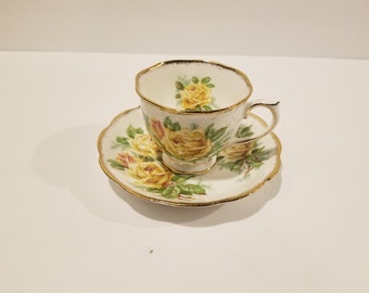 May include: A white teacup and saucer with a gold rim and a floral pattern of yellow roses.